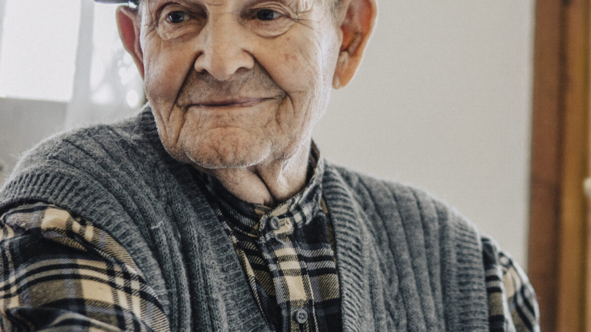 Older man in a flat cap, checked shirt and knitted grey vest smiling gently at home