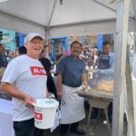 Smiling volunteer in an ALONE T-shirt holds a collection bucket at a street food stall as chefs cook nearby