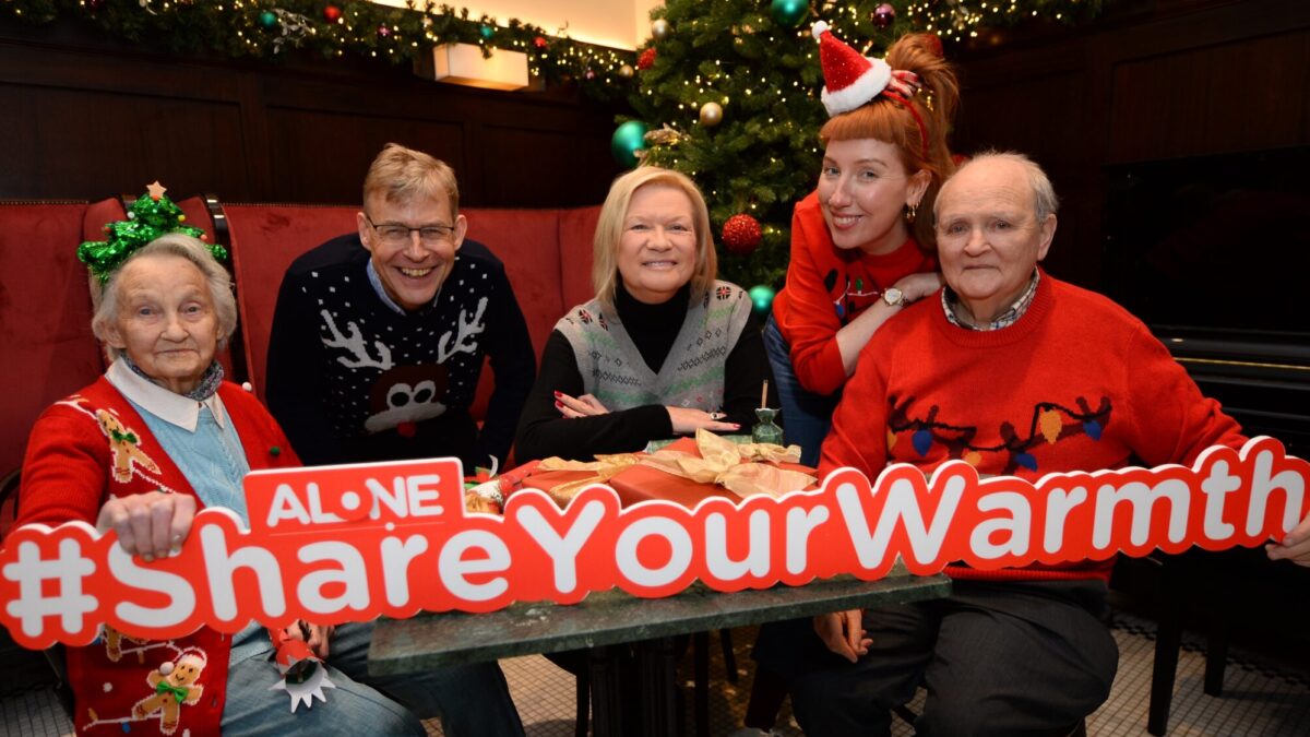 Five people in Christmas jumpers hold a large ALONE '#ShareYourWarmth' sign beside a Christmas tree.
