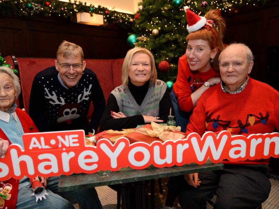 Five people in Christmas jumpers hold a large ALONE '#ShareYourWarmth' sign beside a Christmas tree.