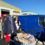 Volunteer in an ALONE T-shirt at an outdoor charity fundraising stall with books, clothes rails and china