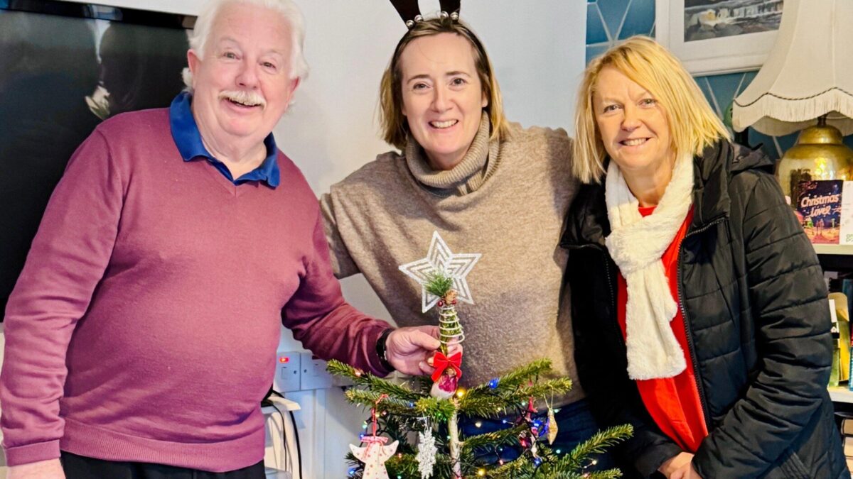An older man smiles beside two women wearing reindeer antlers and a scarf, behind a decorated Christmas tree