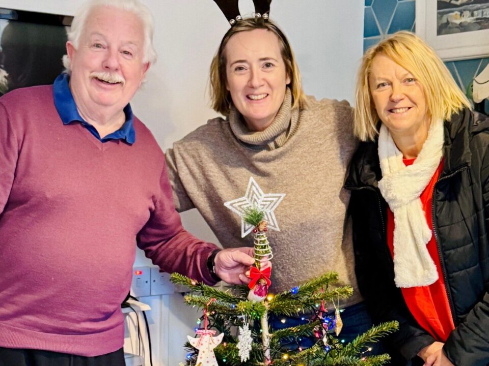 An older man smiles beside two women wearing reindeer antlers and a scarf, behind a decorated Christmas tree