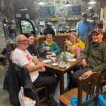 Group of five walkers in outdoor gear smiling around a cafe table with tea, coffee and sandwiches