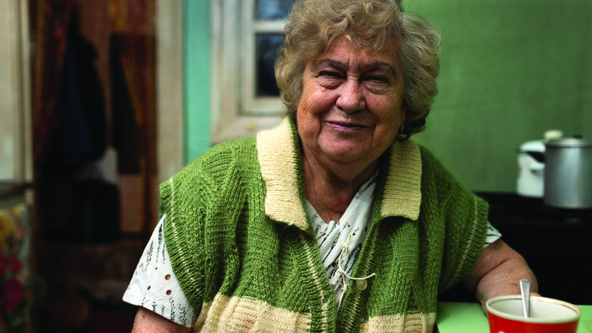 Smiling older woman in a green knitted cardigan sitting at her kitchen table with a mug of tea