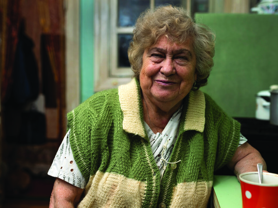 Smiling older woman in a green knitted cardigan sitting at her kitchen table with a mug of tea