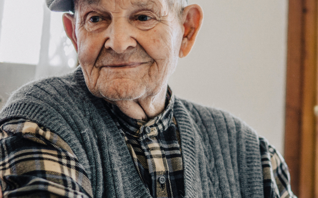 Older man in a flat cap, grey knitted vest and checked shirt smiling gently at home