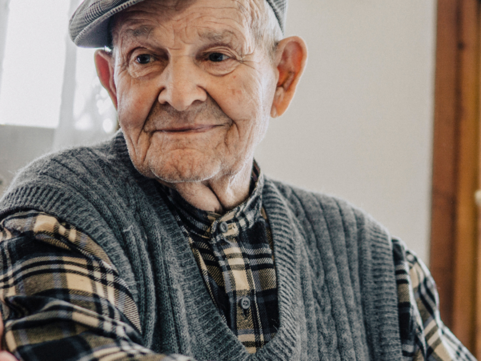Older man in a flat cap, grey knitted vest and checked shirt smiling gently at home