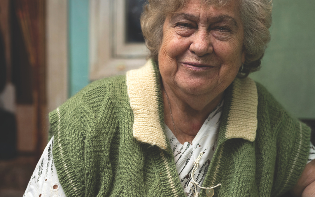 Older woman in a green and cream knitted waistcoat smiling warmly while seated at home