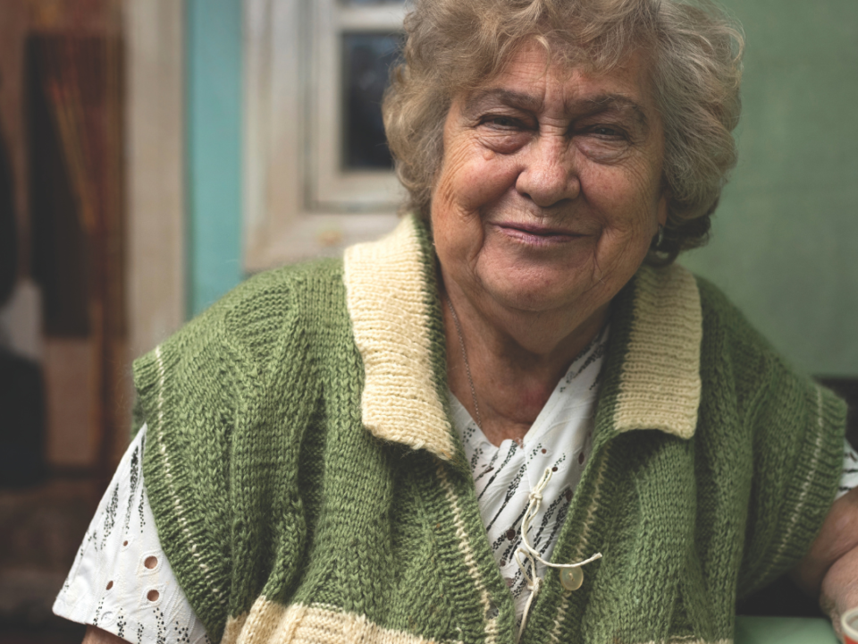 Older woman in a green and cream knitted waistcoat smiling warmly while seated at home