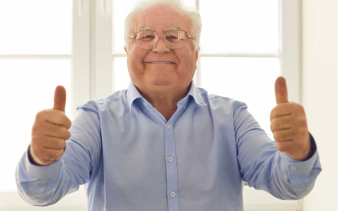 Smiling older man in a blue shirt giving two thumbs up in a bright, sunlit room