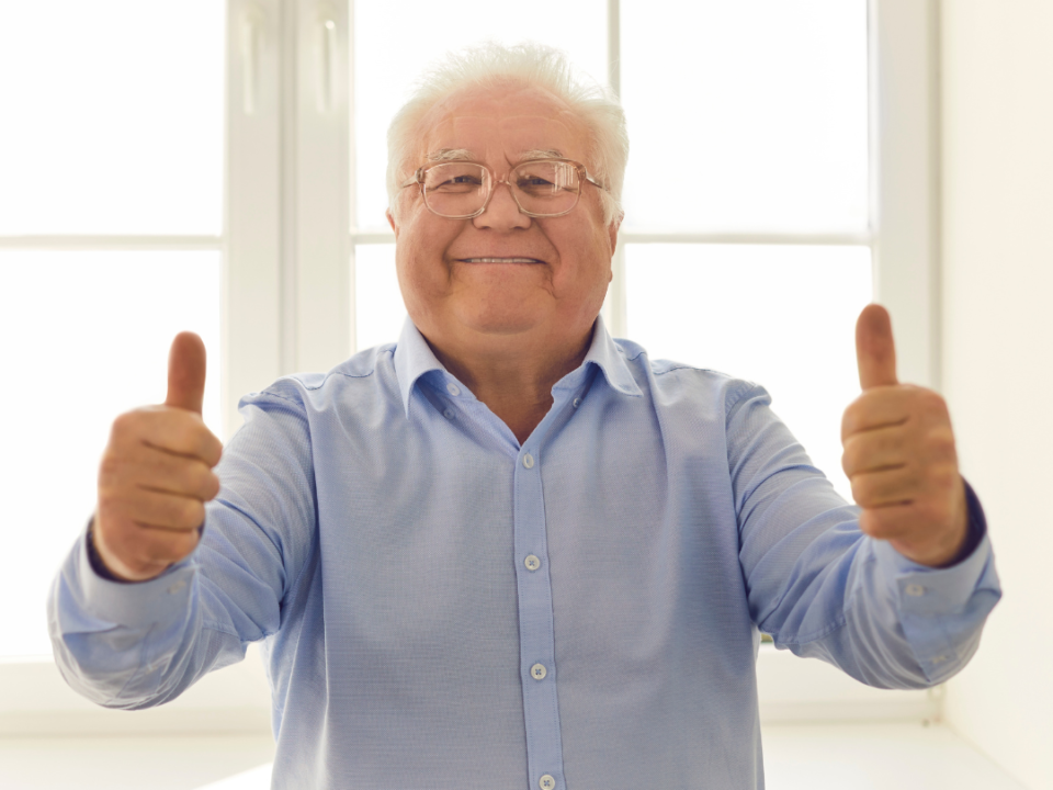 Smiling older man in a blue shirt giving two thumbs up in a bright, sunlit room