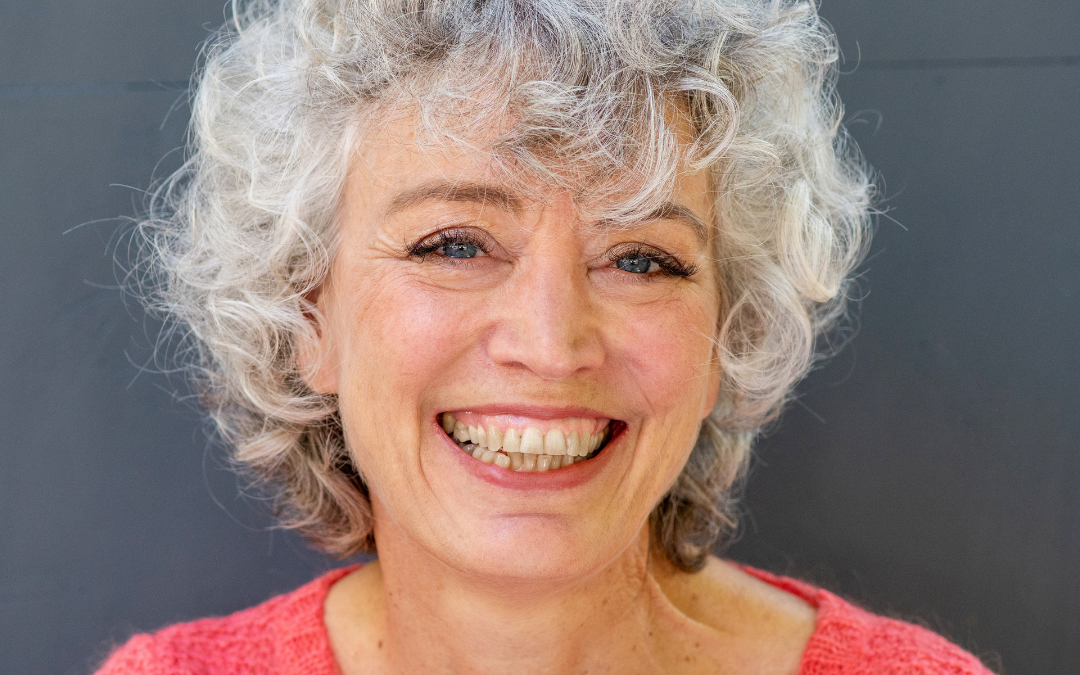 Smiling woman with short curly grey hair wearing a coral knit jumper, against a grey wall