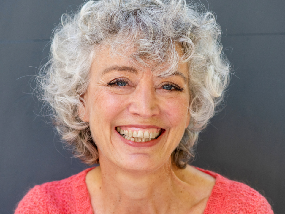 Smiling woman with short curly grey hair wearing a coral knit jumper, against a grey wall