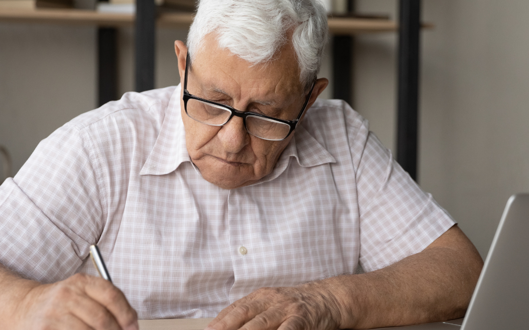 Older man in glasses filling in a form at a desk with a calculator and laptop beside him