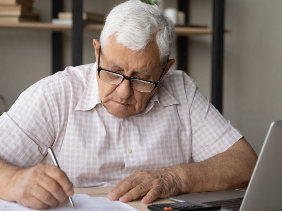Older man in glasses filling in a form at a desk with a calculator and laptop beside him