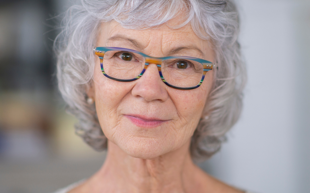 Older woman with short grey curly hair and colourful striped glasses looking calmly at the camera