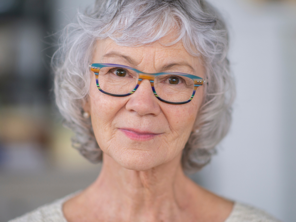 Older woman with short grey curly hair and colourful striped glasses looking calmly at the camera