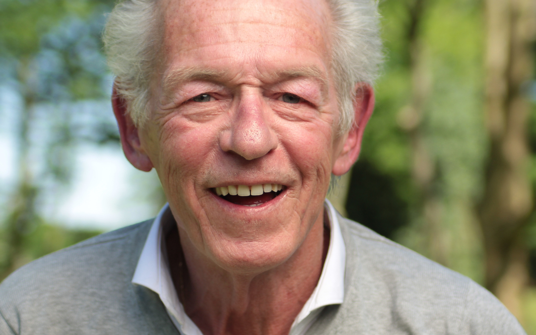 Older man with grey hair smiling warmly outdoors, wearing a grey jumper over a white shirt
