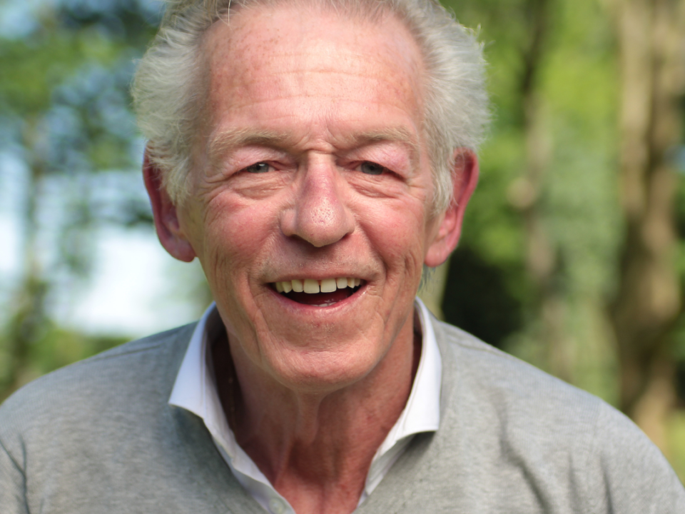 Older man with grey hair smiling warmly outdoors, wearing a grey jumper over a white shirt