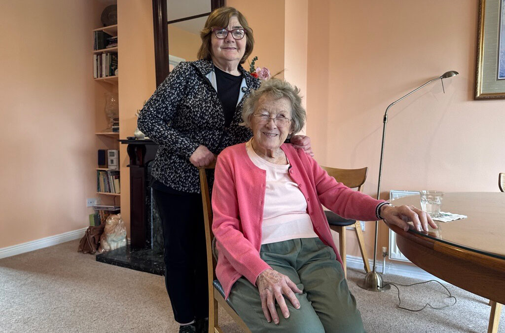 Marian standing beside Bridget, who is seated in her sitting room, both smiling warmly