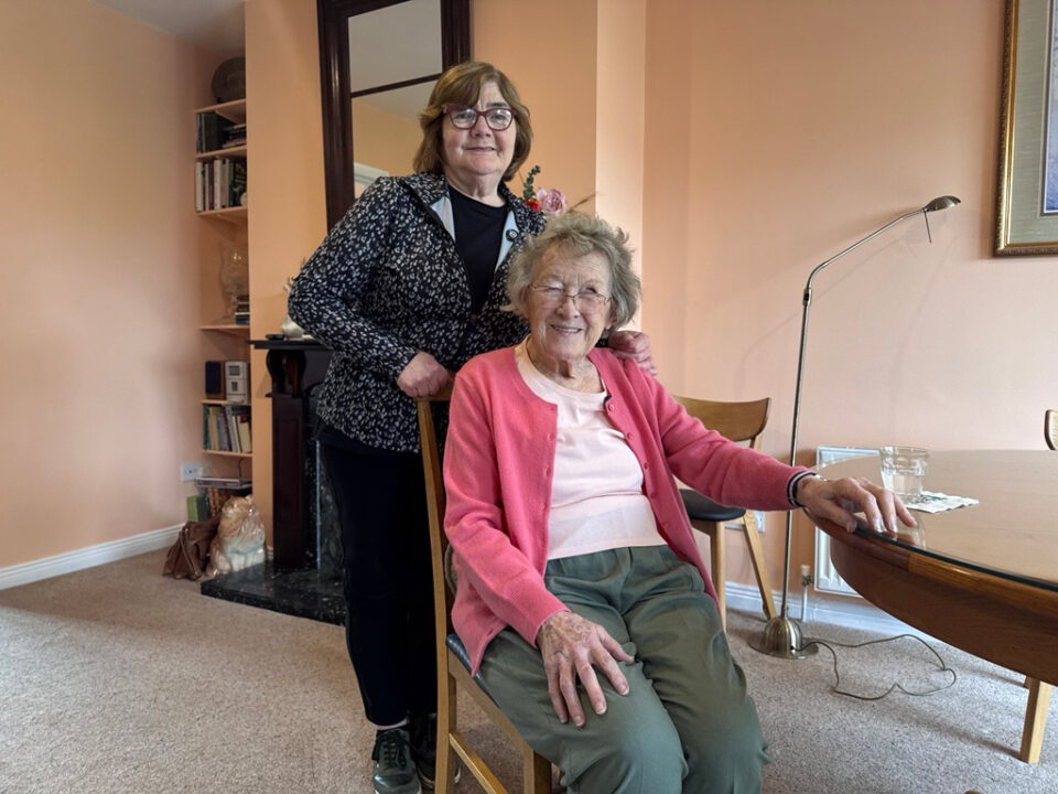 Marian standing beside Bridget, who is seated in her sitting room, both smiling warmly