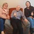 An older man in an armchair gives a thumbs up, with two women seated smiling on either side of him