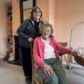 Marian standing beside Bridget, an older woman seated in her sitting room, both smiling warmly