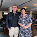 A man in a black jacket stands smiling beside an ALONE staff member wearing a lanyard, in a hotel lounge