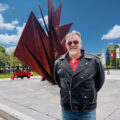 Peter, a smiling man with grey hair, sunglasses and a black leather jacket, standing in a city square