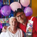An older woman smiling beside a man in a red polo shirt and birthday sash at her 85th birthday party
