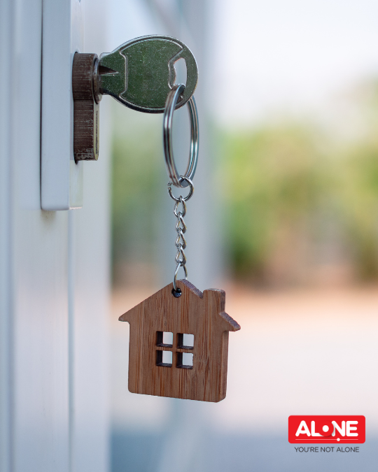 Key in a front door lock with a wooden house-shaped keyring, with ALONE logo 'You're not alone'