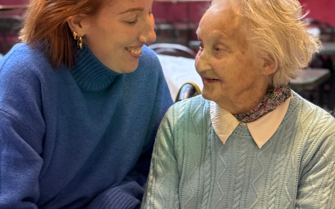 A smiling young woman in a blue jumper holds hands with an older woman in a pale green cable-knit jumper