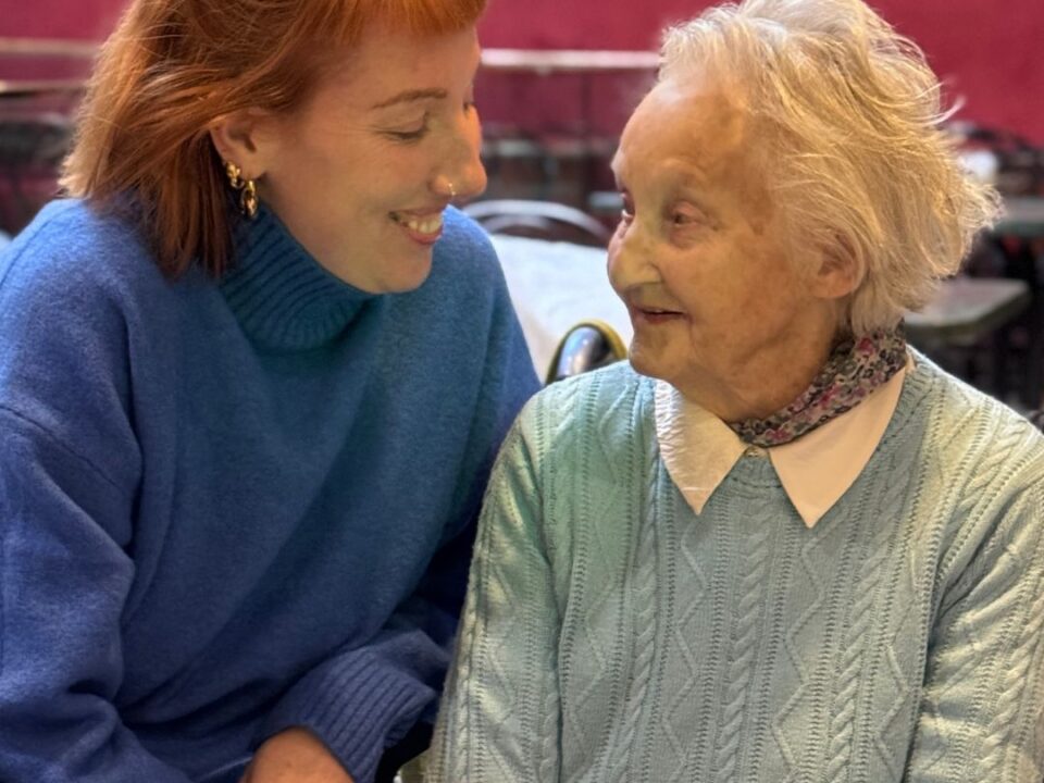A smiling young woman in a blue jumper holds hands with an older woman in a pale green cable-knit jumper