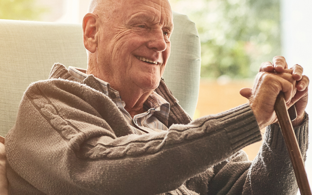 Smiling older man sitting in an armchair at home, resting his hands on a walking stick, with ALONE logo