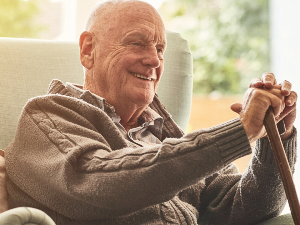 Smiling older man sitting in an armchair at home, resting his hands on a walking stick, with ALONE logo