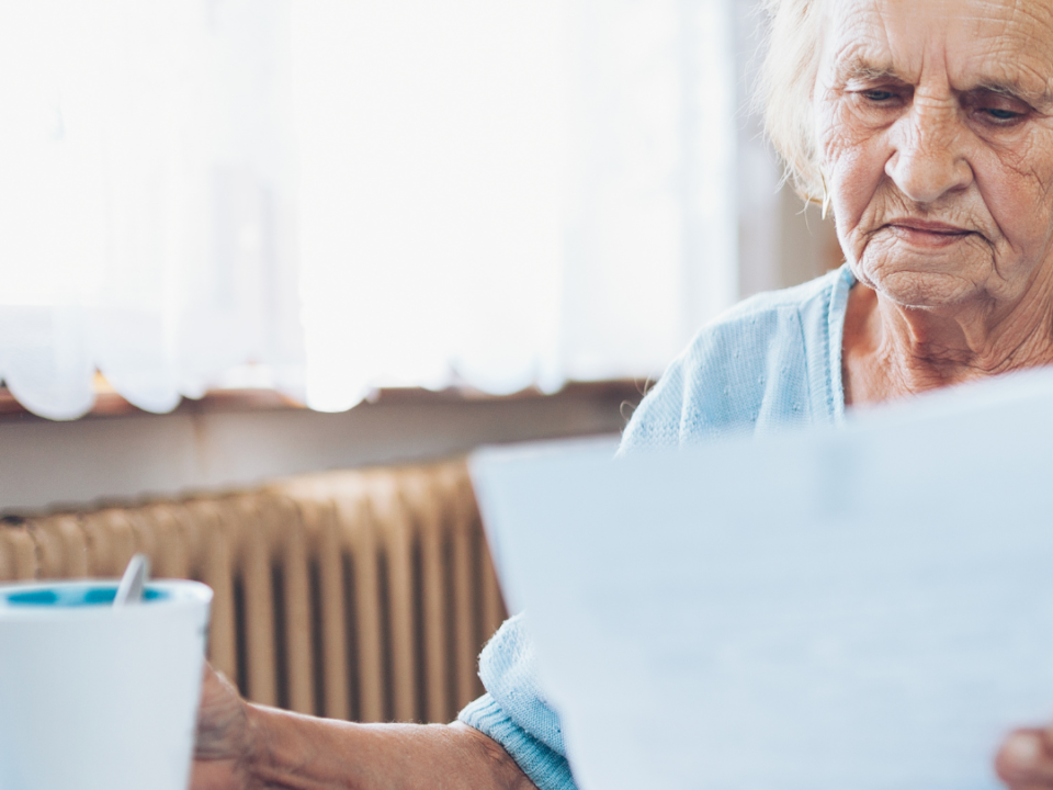 Older woman at a table with a cup of tea, reading paperwork with a concerned expression