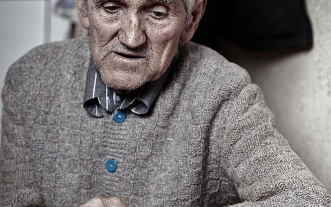 Older man in a heavy knitted cardigan sitting with hands clasped at a table at home, looking downward