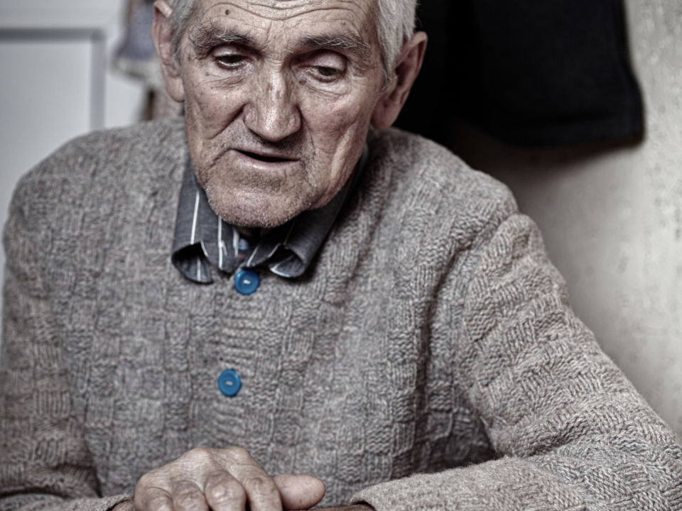 Older man in a heavy knitted cardigan sitting with hands clasped at a table at home, looking downward