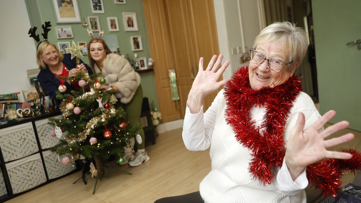 Older woman with red tinsel around her neck laughs while two others decorate a Christmas tree behind her