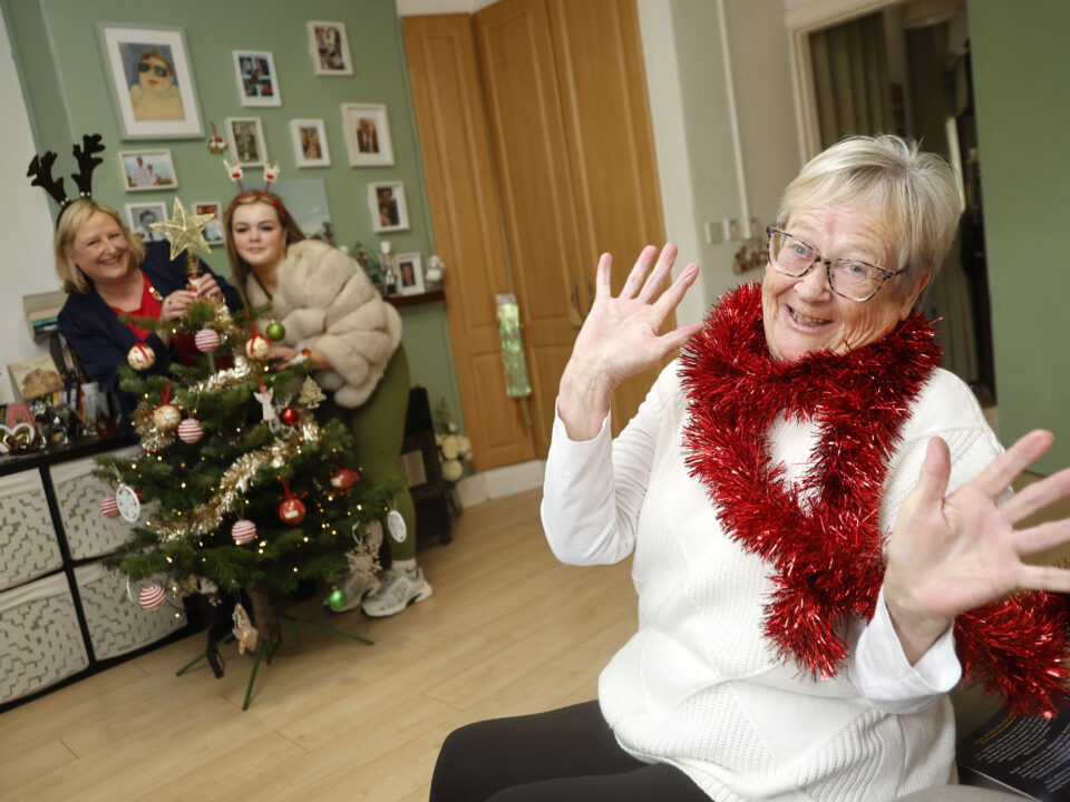 Older woman with red tinsel around her neck laughs while two others decorate a Christmas tree behind her