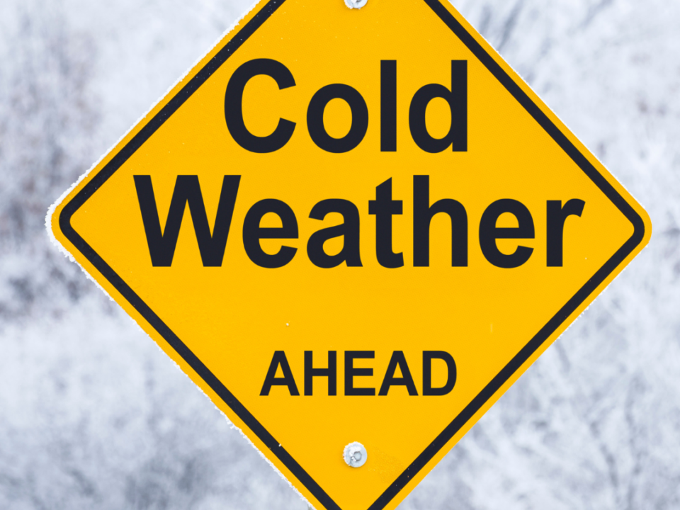 Yellow diamond road sign reading "Cold Weather Ahead" against a frosty, snow-covered landscape