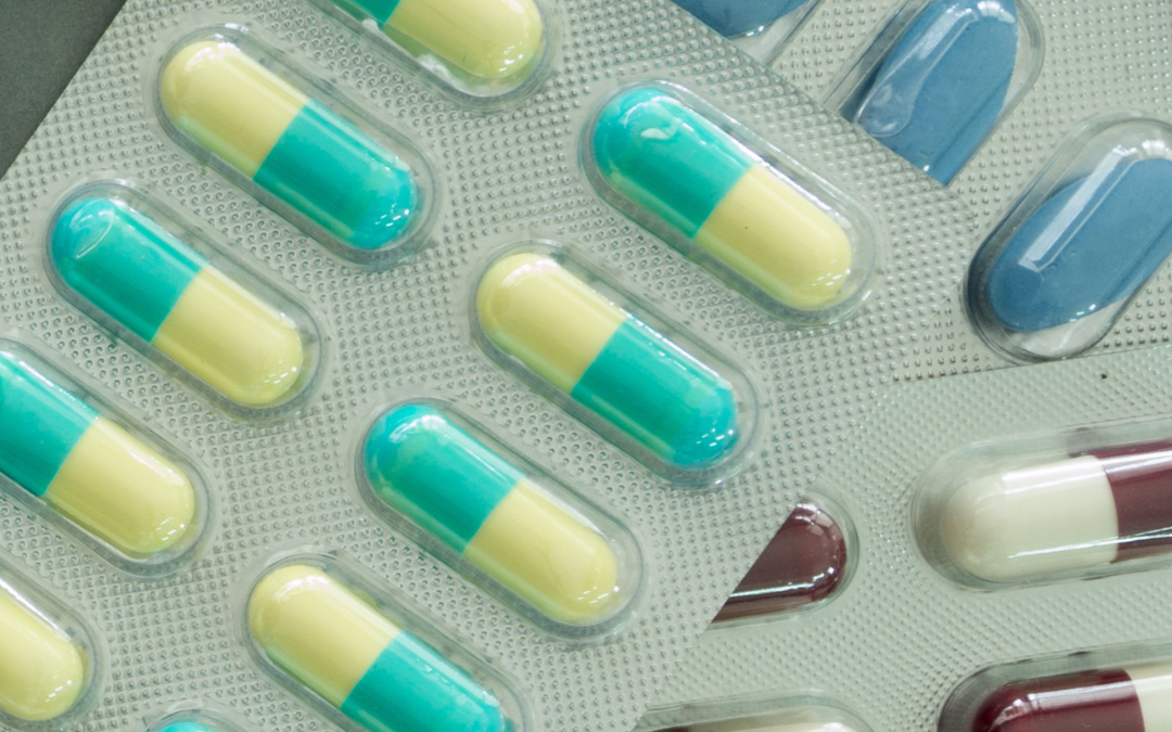 Blister packs of green, yellow, blue and maroon capsules and tablets overlapping on a grey surface