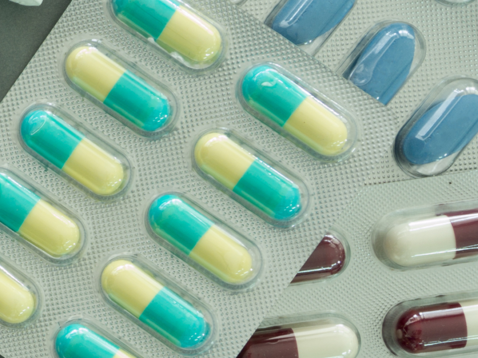 Blister packs of green, yellow, blue and maroon capsules and tablets overlapping on a grey surface