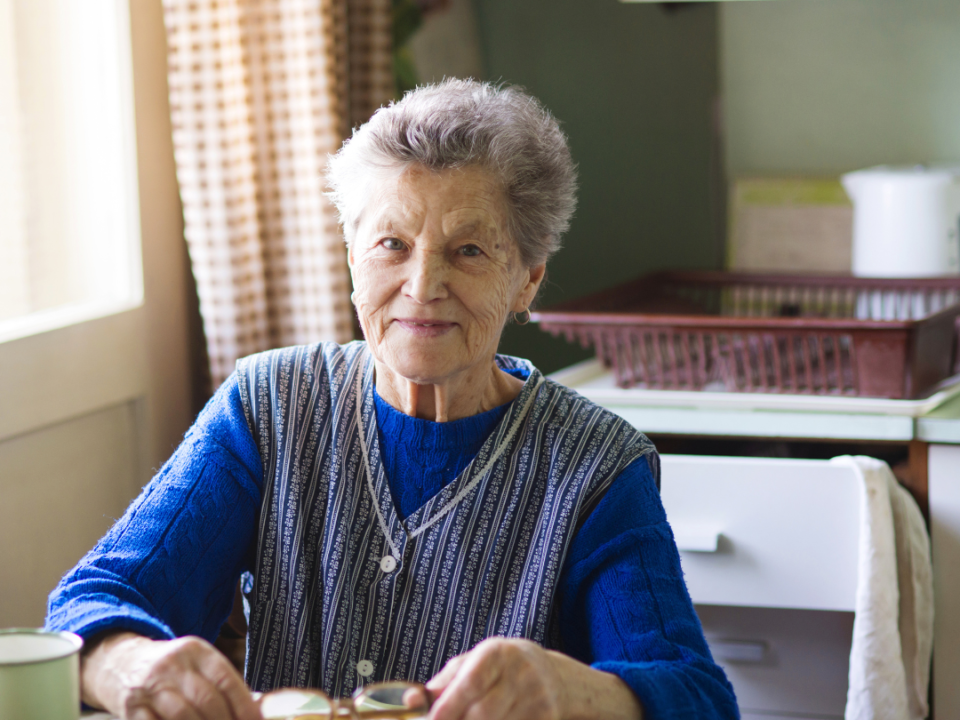 Older woman sitting at her kitchen table at home, smiling, with glasses and a cup in front of her