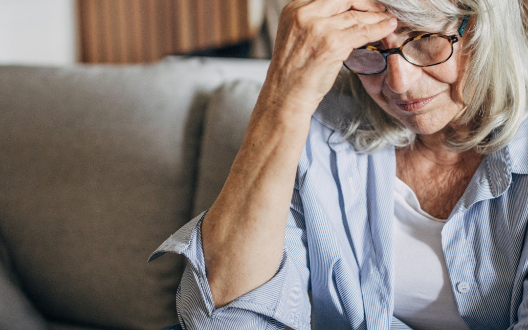 An older woman sits on a sofa at home, resting her hand on her forehead with a worried expression