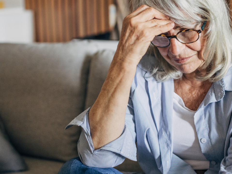 An older woman sits on a sofa at home, resting her hand on her forehead with a worried expression