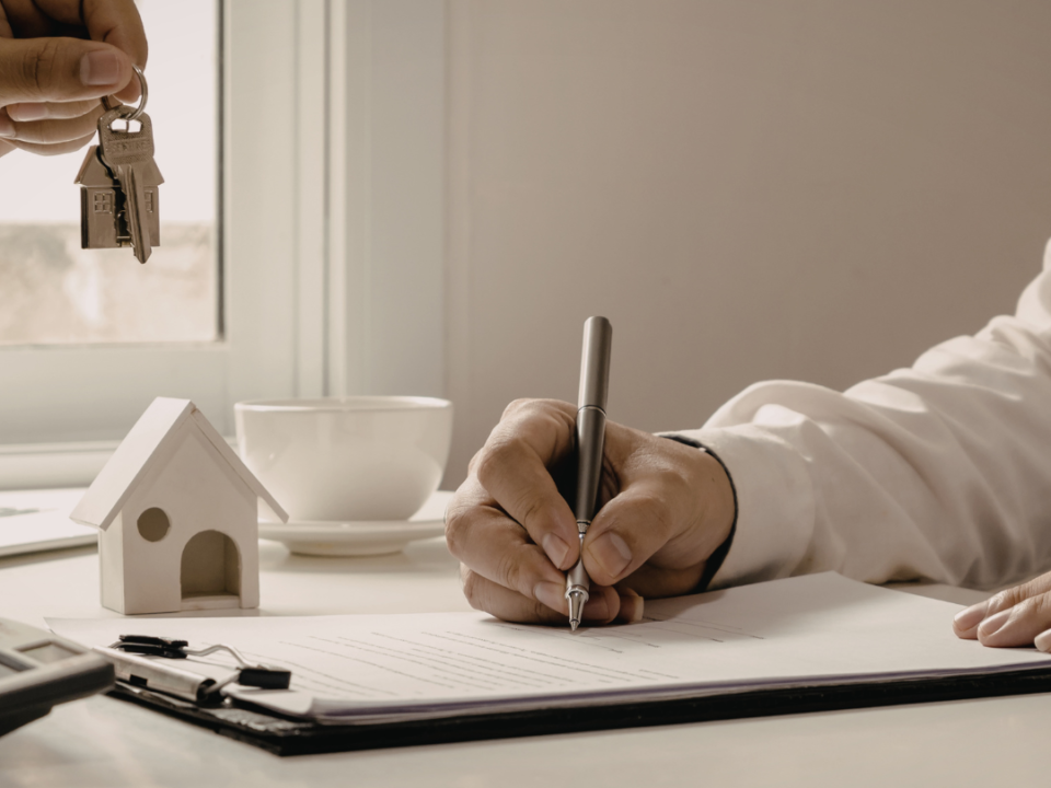A person signing housing paperwork at a desk while another hand holds out house keys