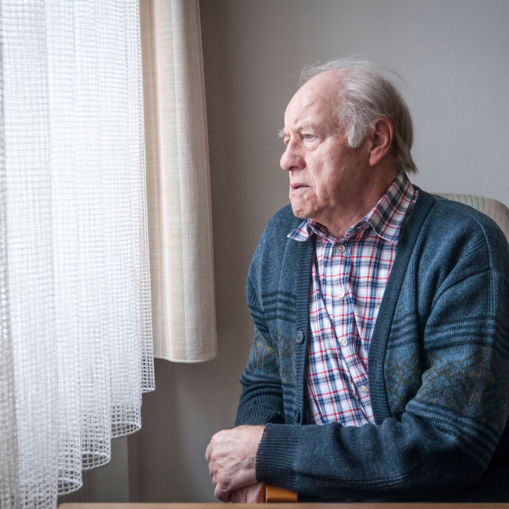 An older man in a blue cardigan and checked shirt sits at home, looking out through a net-curtained window.