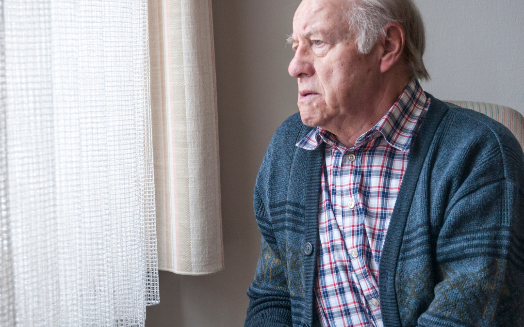 An older man in a blue cardigan and checked shirt sits at home, looking out through a net-curtained window.